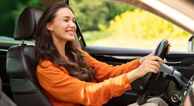 Young woman with long brown hair and a long sleeve orange shirt with both hands on the wheel driving her car