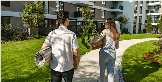 A man with a rug rolled up under his arm walks with a toman carrying a basket in a condo complex